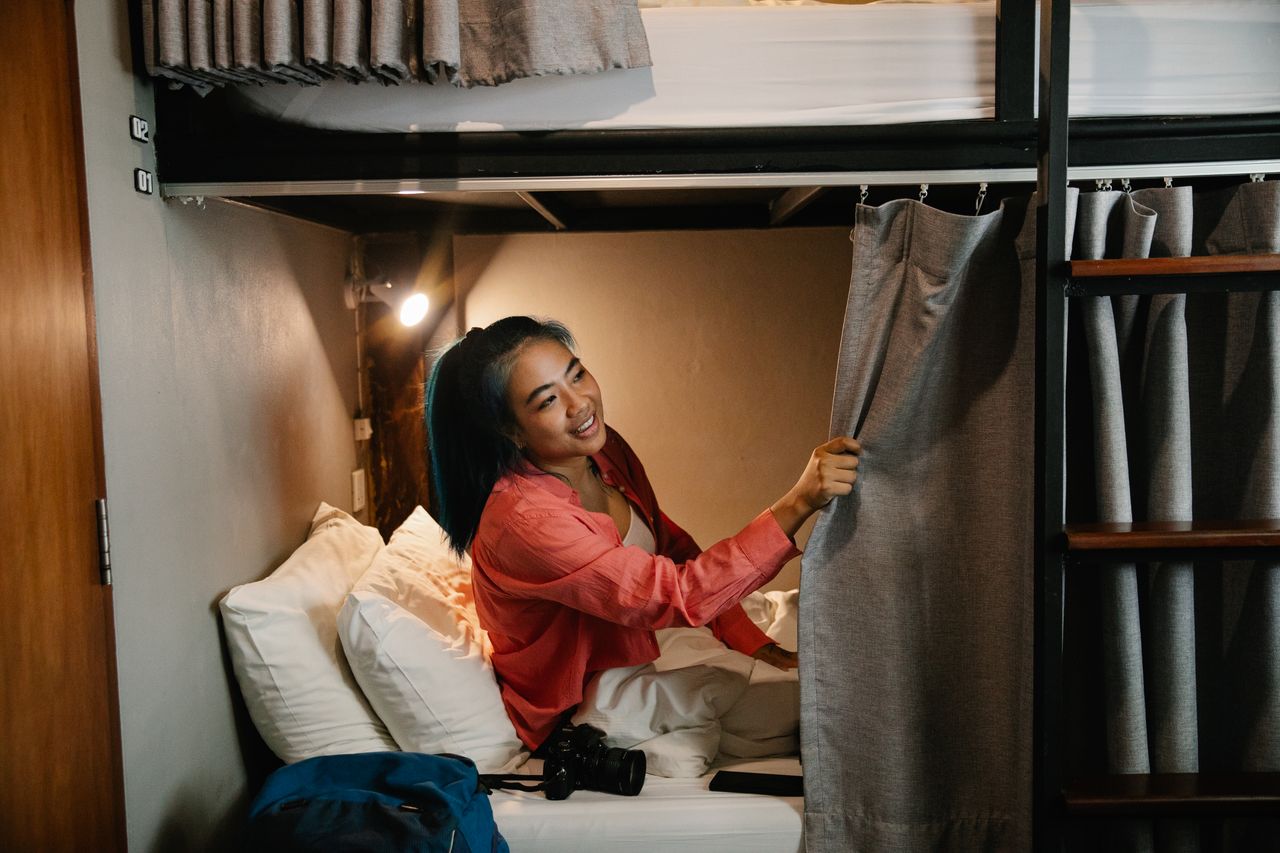 Side view of young smiling ethnic female resting in bed under white blanket with camera and phone and looking away holding curtain in train